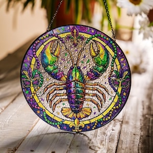 Op de afbeelding: Rond gebrandschilderd glazen raam met een kleurrijk kreeftontwerp. De kreeft is afgebeeld in paars, groen en geel, omringd door Mardi Gras-thema-accenten. Het raam heeft een zwarte rand en een ophangketting.