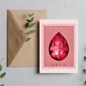 May include: A greeting card featuring a ruby gemstone illustration. The card has a pink background with the words "RUBY" and "JULY" printed on it. A brown envelope and a sprig of greenery are next to the card.