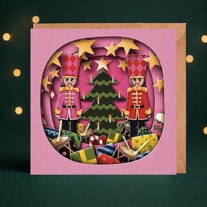 May include: A square greeting card featuring a layered paper-cut design with a circular cutout. The scene displays two nutcracker figures in pink and red uniforms flanking a green Christmas tree, surrounded by yellow stars and wrapped gifts.