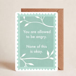 May include: A rectangular greeting card with a scalloped edge, featuring a light green background and white line art of leafy branches. The text reads: You are allowed to be angry. None of this is okay. A brown envelope is positioned behind the card.