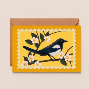 May include: A rectangular greeting card featuring a black and white magpie perched on a branch with white flowers against a bright yellow background, designed with a decorative scalloped border resembling a postage stamp, paired with a brown envelope.