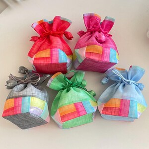 May include: Six colorful, small fabric pouches with drawstring closures. The pouches are in shades of red, pink, green, blue, and gray, with patchwork designs in various colors. The drawstring ties are in matching or complementary colors.