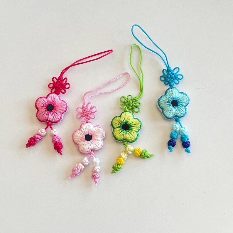 Pu&ograve; includere: Quattro ornamenti floreali ricamati colorati. Ognuno ha un design floreale con un centro a contrasto, un anello per appendere e nappe decorative. I colori includono rosa, rosa chiaro, verde e blu.