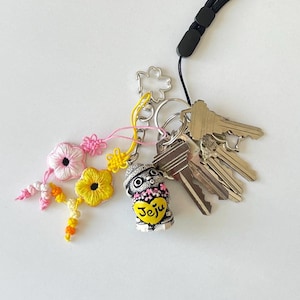 Pu&ograve; includere: Un portachiavi con una piccola statuetta decorativa a forma di cane con un cappello e un cuore con la scritta "Jeju". Il portachiavi include anche un mazzo di chiavi, un ciondolo floreale e un cordino nero.