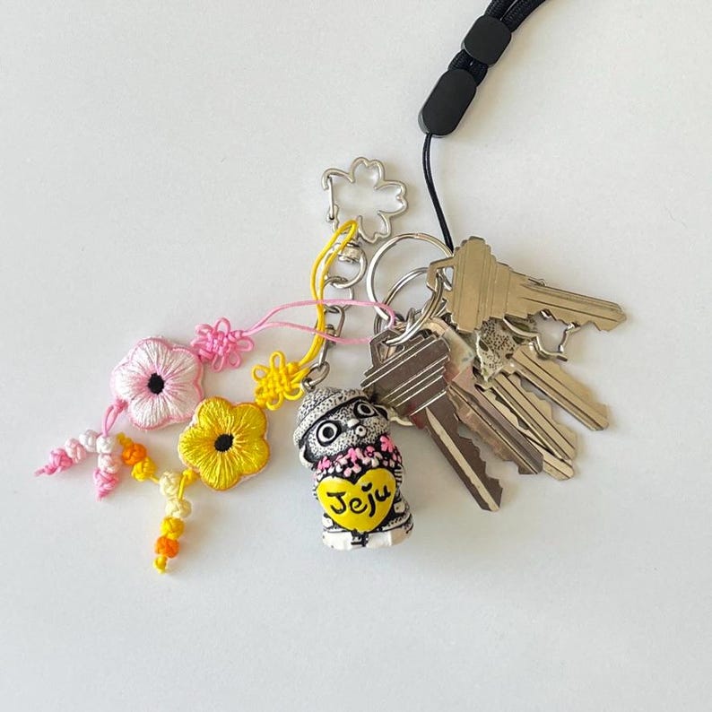 Pu&ograve; includere: Un portachiavi con una piccola statuetta decorativa a forma di cane con un cappello e un cuore con la scritta "Jeju". Il portachiavi include anche un mazzo di chiavi, un ciondolo floreale e un cordino nero.