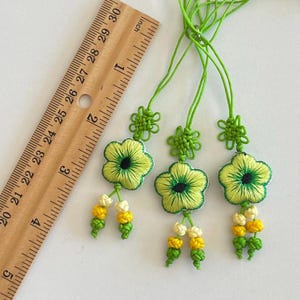 Pu&ograve; includere: Tre ornamenti floreali ricamati con dettagli verdi e gialli. Ogni ornamento presenta una forma di fiore con un centro verde e un nappa di perline gialle e bianche. Gli ornamenti sono attaccati a cordoncini verdi.
