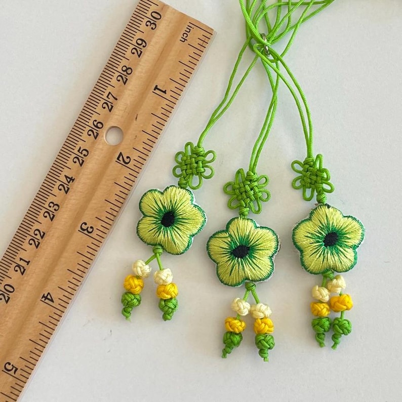 Pu&ograve; includere: Tre ornamenti floreali ricamati con dettagli verdi e gialli. Ogni ornamento presenta una forma di fiore con un centro verde e un nappa di perline gialle e bianche. Gli ornamenti sono attaccati a cordoncini verdi.