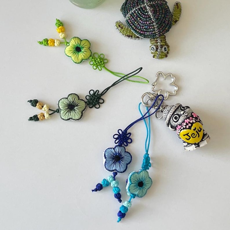 Pu&ograve; includere: Un assortimento di portachiavi e ciondoli fatti a mano. L'immagine presenta una tartaruga di perline, accenti floreali ricamati in verde e blu e una piccola statuina a tema Jeju. I portachiavi hanno vari colori e design.