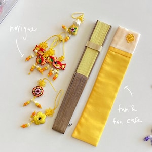 Pu&ograve; includere: Un assortimento di accessori tradizionali coreani, tra cui un ventaglio giallo, una custodia per ventaglio e ornamenti decorativi norigae. Il ventaglio &egrave; marrone e giallo, e la custodia &egrave; gialla con una parte superiore color crema. I norigae presentano ricami colorati.