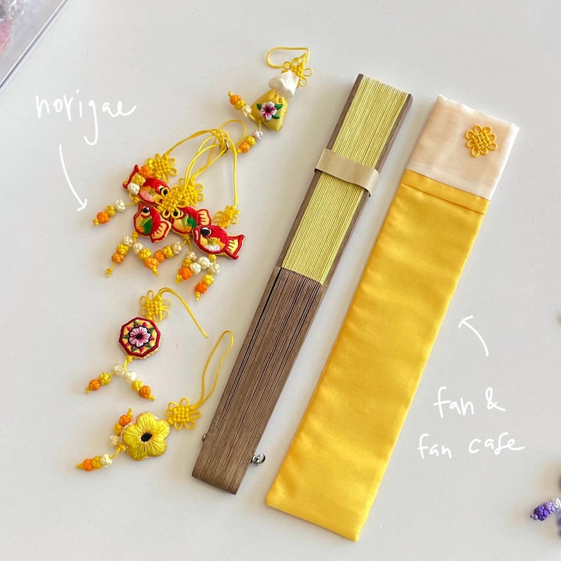 Pu&ograve; includere: Un assortimento di accessori tradizionali coreani, tra cui un ventaglio giallo, una custodia per ventaglio e ornamenti decorativi norigae. Il ventaglio &egrave; marrone e giallo, e la custodia &egrave; gialla con una parte superiore color crema. I norigae presentano ricami colorati.
