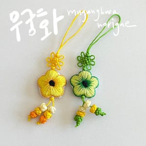Pu&ograve; includere: Due ornamenti floreali ricamati. Uno &egrave; giallo con un centro nero e l'altro &egrave; verde con un centro verde scuro. Entrambi hanno un anello per appenderli e nappe decorative. L'immagine ha testo coreano e la parola "norigne".