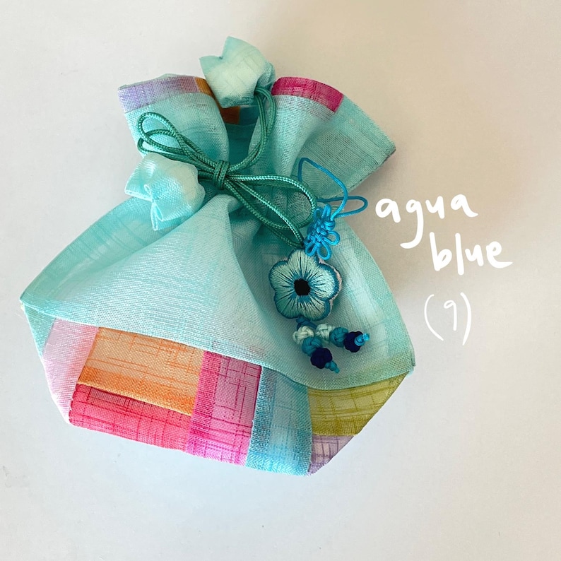Pu&ograve; includere: Pochette in tessuto blu acqua con chiusura a coulisse e un ciondolo floreale decorativo. La pochette presenta un design patchwork con pannelli rosa, arancioni e verdi. Il testo "aqua blue (9)" &egrave; scritto sul lato.
