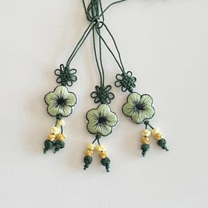 Pu&ograve; includere: Tre ornamenti floreali fatti a mano con dettagli ricamati verdi e neri. Ogni ornamento presenta un fiore verde chiaro con un centro scuro, un nodo decorativo e perline gialle e verdi pendenti. Gli ornamenti sono sospesi da cordoncini verdi.