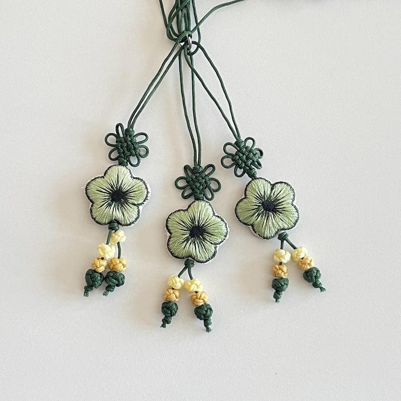 Pu&ograve; includere: Tre ornamenti floreali fatti a mano con dettagli ricamati verdi e neri. Ogni ornamento presenta un fiore verde chiaro con un centro scuro, un nodo decorativo e perline gialle e verdi pendenti. Gli ornamenti sono sospesi da cordoncini verdi.