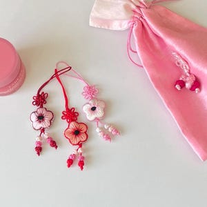 Pu&ograve; includere: Ornamenti floreali ricamati in tonalit&agrave; rosa e rosse, con intricati dettagli di nodi. Sono visibili anche un sacchetto rosa con coulisse e un vasetto di balsamo per le labbra rosa. Gli ornamenti hanno un aspetto delicato e artigianale.