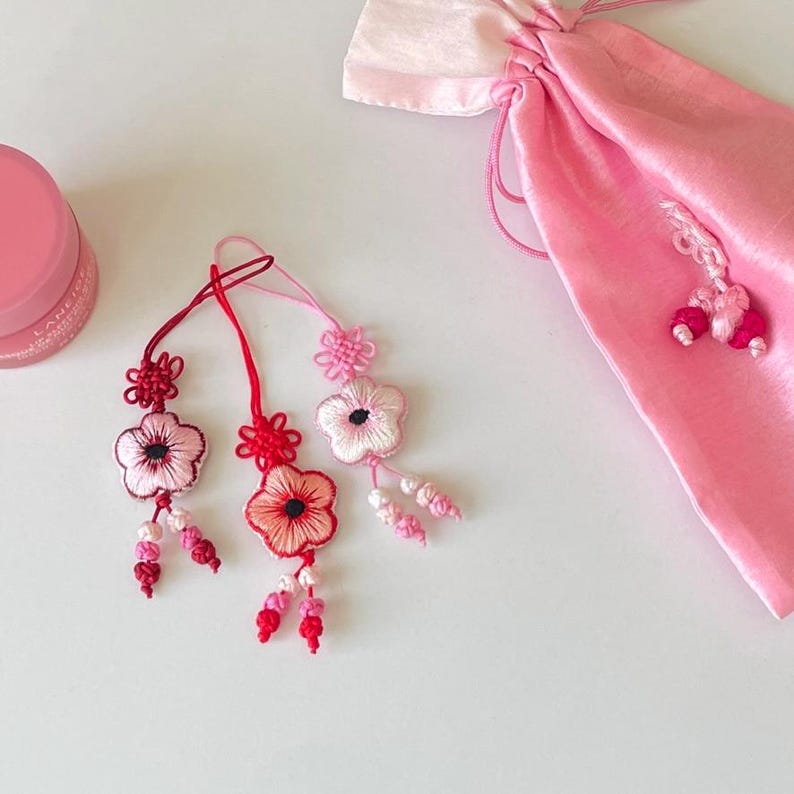 Pu&ograve; includere: Ornamenti floreali ricamati in tonalit&agrave; rosa e rosse, con intricati dettagli di nodi. Sono visibili anche un sacchetto rosa con coulisse e un vasetto di balsamo per le labbra rosa. Gli ornamenti hanno un aspetto delicato e artigianale.