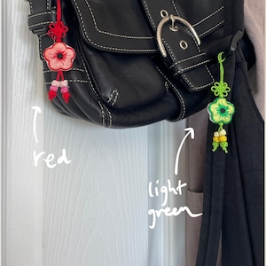 Pu&ograve; includere: Una borsa in pelle nera con fibbia argentata e cuciture bianche. Due ornamenti floreali decorativi sono appesi alla borsa. Uno &egrave; rosso con accenti rosa, l'altro &egrave; verde chiaro con accenti gialli.