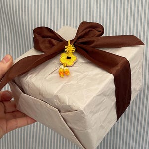 Pu&ograve; includere: Un pacco regalo avvolto con un nastro di raso marrone e un ornamento floreale giallo. Il regalo &egrave; avvolto in carta colorata. Lo sfondo &egrave; un motivo a righe blu chiaro e bianco.