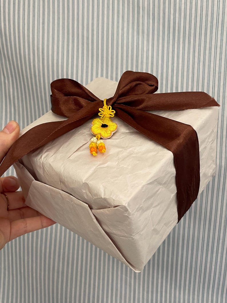 Pu&ograve; includere: Un pacco regalo avvolto con un nastro di raso marrone e un ornamento floreale giallo. Il regalo &egrave; avvolto in carta colorata. Lo sfondo &egrave; un motivo a righe blu chiaro e bianco.