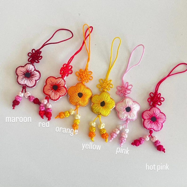 Pu&ograve; includere: Una collezione di sei ciondoli floreali ricamati in vari colori: bordeaux, rosso, arancione, giallo, rosa e rosa acceso. Ogni ciondolo presenta un motivo floreale, un anello per appendere e un nappa di perline. I colori sono etichettati.