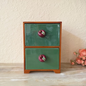 May include: A small wooden storage chest featuring two stacked drawers with glossy green ceramic faces. Each drawer is equipped with a singular knob shaped like a small red apple with a green leaf detail, providing a functional and decorative accent.