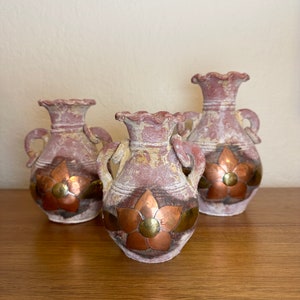 Puede incluir: Tres jarrones de color terracota con detalles de flores de cobre. Cada jarrón presenta un borde con volantes, dos asas y un acabado envejecido. Las flores de cobre tienen un botón central de latón.