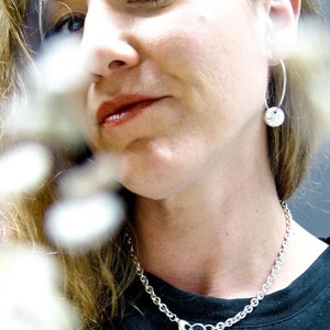 Peut inclure: Une femme portant un collier en chaîne argent avec un pendentif rond et des boucles d'oreilles créoles argent avec un pendentif rond. Le collier et les boucles d'oreilles ont une texture martelée.