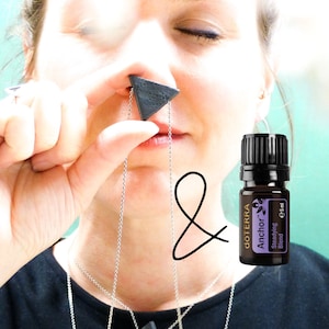 Op de afbeelding: Een vrouw draagt een zwart shirt met twee zilveren kettingen, een met een zwarte driehoekige hanger. Ze houdt een klein flesje etherische olie vast met het label "dōTERRA Anchor™ Sleepifying Blend 5 ml".