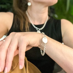 Może przedstawiać: Srebrny naszyjnik z łańcuszkiem i okrągłym wisiorem oraz srebrna bransoletka z łańcuszkiem i okrągłym wisiorem. Zarówno naszyjnik, jak i bransoletka są noszone przez osobę w czarnym topie na ramiączkach.