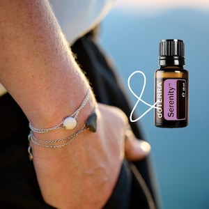 May include: A silver bracelet with a white lava bead and a small black charm. A bottle of doTERRA Serenity essential oil is shown in the background.