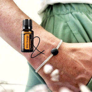 May include: A brown glass bottle of Wild Orange essential oil from doTERRA. The bottle is labelled with the brand name and the name of the oil. The bottle is sitting on a person's wrist, which is wearing a silver bracelet with a black lava rock bead.