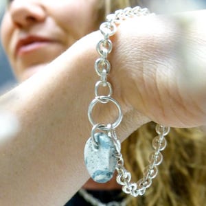 Può includere: Un braccialetto a catena d'argento con un grande ciondolo ovale. Il ciondolo è realizzato in un materiale grigio chiaro con venature grigio scuro. Il braccialetto è al polso di una persona.