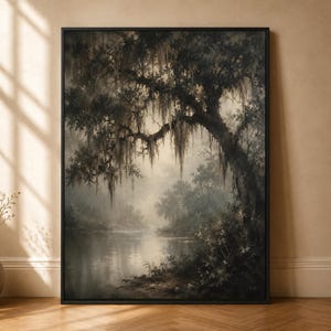 Op de afbeelding: Een ingelijste muurkunstprint met een donker, sfeervol landschap van een rivieroever met een grote, uitgestrekte boom bedekt met hangend mos. De scène bevat gedempte tinten houtskool, diepgroen en zacht beige voor een mistig effect.