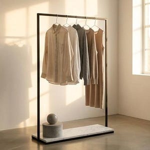 Pode incluir: Um cabideiro de metal preto com base de mármore branco, segurando quatro peças de roupa em cabides: uma camisa bege, uma camisa branca, um suéter cinza e um vestido sem mangas bege. Um objeto cilíndrico de pedra cinza repousa na base.