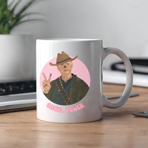 Può includere: Tazza in ceramica bianca con un design circolare rosa che presenta un personaggio dei cartoni animati con un cappello da cowboy e che fa il segno della pace. Il testo "Ghoul Power" è scritto in rosa sotto il personaggio.