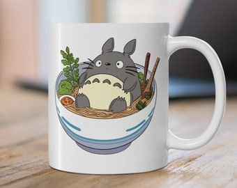 May include: White ceramic mug with a cartoon illustration of a gray Totoro character sitting in a bowl of ramen noodles. The bowl has blue and white stripes and there are chopsticks and green vegetables in the bowl.
