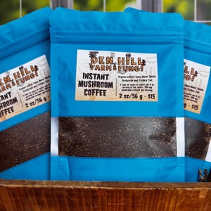Può includere: Una bustina blu con un'etichetta bianca che recita "Den Hill Farm & Fungi Instant Mushroom Coffee 2 oz/56 g - $15". La bustina contiene una polvere marrone scuro. L'etichetta elenca anche gli ingredienti: caffè biologico, criniera di leone, reishi, cordyceps e coda di tacchino.
