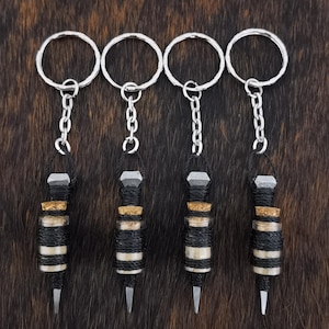 Puede incluir: Cuatro llaveros con anillas y cadenas plateadas. Cada llavero presenta un pequeño diseño cilíndrico envuelto en hilo blanco y negro, con un tapón de corcho y una punta plateada. El fondo es una superficie marrón texturizada.