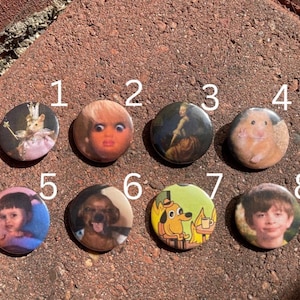 May include: A collection of eight round pinback buttons with various images. The images include a doll, a hamster, a painting, a dog, a child, and a cartoon dog with the text "This is fine".