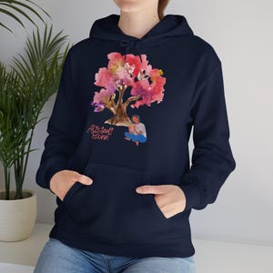 Könnte beinhalten: Ein marineblauer Hoodie mit einem rosa Aquarellbaum und einer Person, die darunter sitzt. Der Text "Altstadt Bonn" ist auf dem Hoodie aufgedruckt.