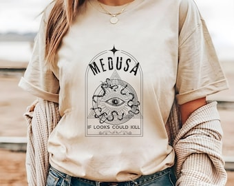 May include: A beige t-shirt with a black graphic print of Medusa's face surrounded by snakes. The text "Medusa" and "If looks could kill" are printed on the shirt.