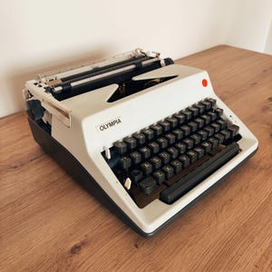 Könnte beinhalten: Eine Olympia Schreibmaschine mit cremefarbenem Gehäuseoberteil und dunkelgrauer Basis. Das Gerät verfügt über eine QWERTY-Tastatur mit schwarzen Tasten, einen Metallwagen und eine kleine rote, kreisförmige Kontrollleuchte oben rechts.