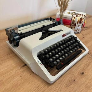 Könnte beinhalten: Eine weiße und dunkelblaue Erika Schreibmaschine mit QWERTY-Tastatur, schwarzen runden Tasten, einem metallenen Wagenrücklaufhebel und einem roten Markenlogo in der oberen rechten Ecke. Das Gehäuse hat ein schlankes, angewinkeltes Design.