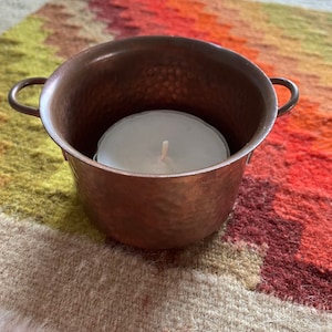 Puede incluir: Un portavelas de cobre martillado con una vela de té blanca en su interior. El portavelas tiene dos asas y está sobre una alfombra multicolor.