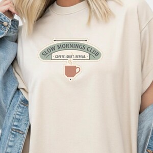 May include: A cream-coloured t-shirt with a graphic that reads "SLOW MORNINGS CLUB" above a coffee cup illustration. The text "COFFEE. QUIET. REPEAT." is below the club name. The shirt has short sleeves and a relaxed fit.