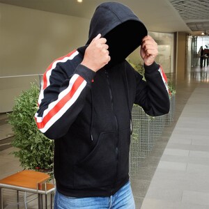 May include: Black zip-up hoodie with red and white stripes on the sleeves. The hoodie has a hood and a front pocket. The person is wearing blue jeans. The background is a hallway with a plant and a table.
