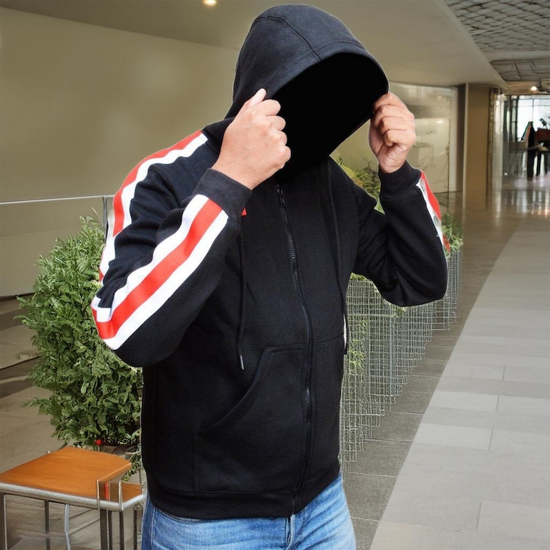 May include: Black zip-up hoodie with red and white stripes on the sleeves. The hoodie has a hood and a front pocket. The person is wearing blue jeans. The background is a hallway with a plant and a table.