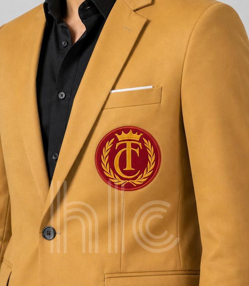 May include: Mustard yellow blazer with a black shirt underneath. The blazer features a red and gold circular emblem on the chest. A white pocket square is visible in the chest pocket. The blazer has a two-button closure.