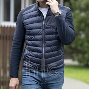 May include: A navy blue quilted puffer jacket with a full-length zipper, paired with dark blue knit sleeves and a stand-up collar. The jacket is worn with blue jeans and a white undershirt.