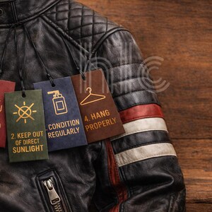 May include: Close-up of a black leather jacket with a quilted shoulder design. Attached tags provide care instructions: avoid water, keep out of direct sunlight, condition regularly, and hang properly. The jacket has a zipper closure and decorative stripes.